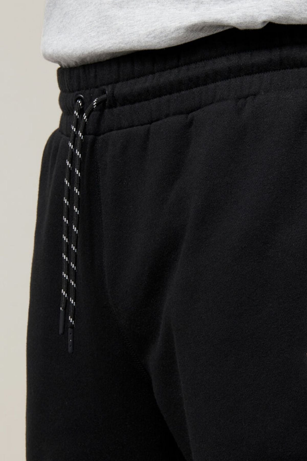 Close-up of a person wearing the Hard Yakka Xtreme Jogger, featuring black track pants with a black and white striped drawstring and reinforced knee patches. The waistband is elastic, and the individual is also sporting a light gray shirt. The background is plain and out of focus.
