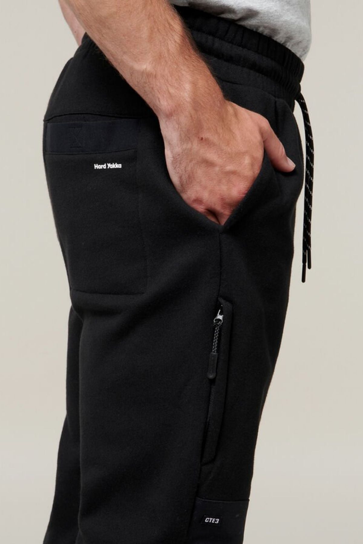 A person wearing Hard Yakka Xtreme Jogger in black with a drawstring waistband. The joggers feature reinforced knee patches, a zippered pocket, and another pocket labeled "Henri Yedder." The person's hand is casually placed in one of the pockets. The person is also wearing a light-colored top.