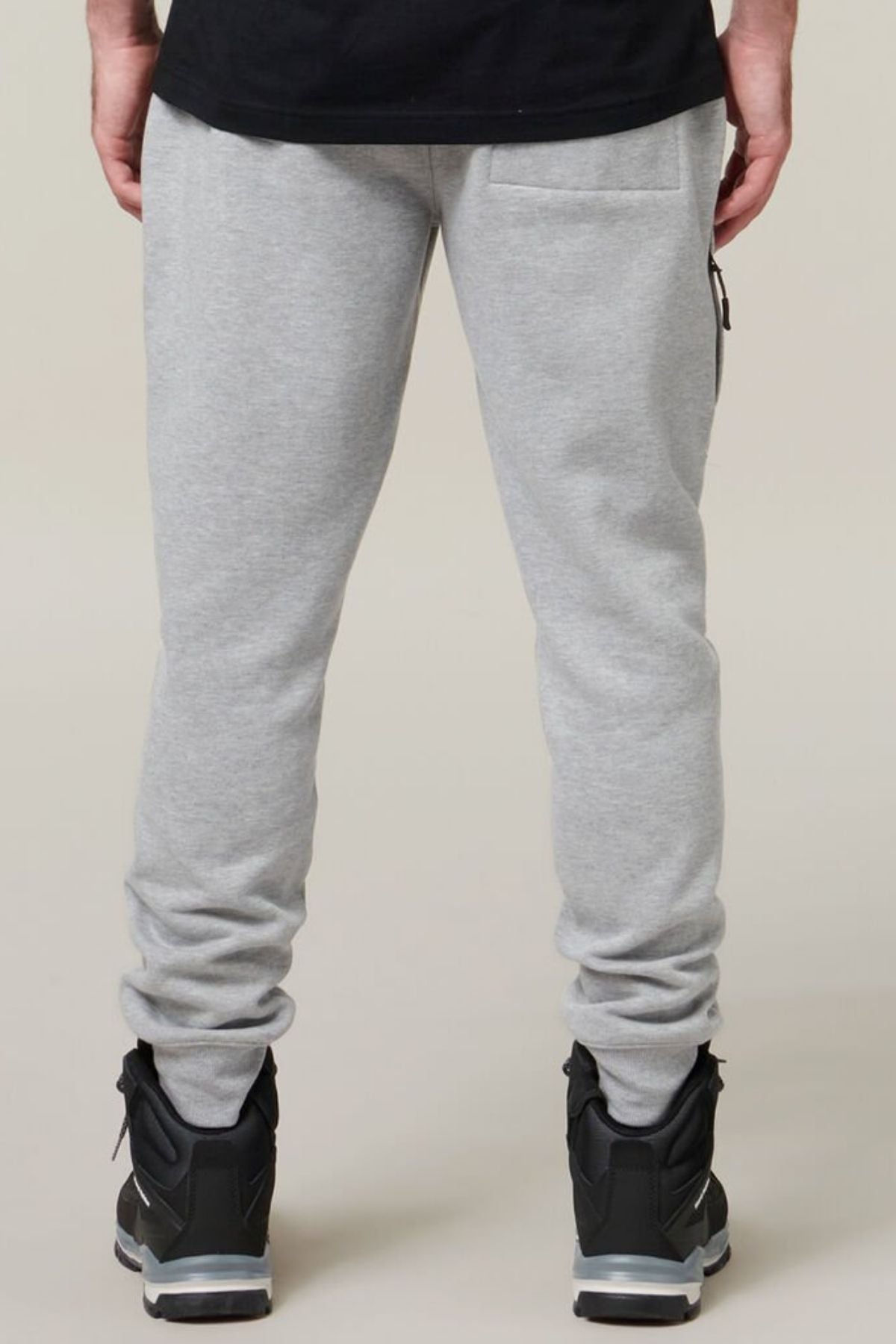A person stands facing away from the camera, wearing Hard Yakka Xtreme Joggers in light gray, featuring reinforced knee patches and a back right side pocket. The joggers are slightly gathered at the ankles and paired with black sneakers that have white soles. The individual is also dressed in a black top.