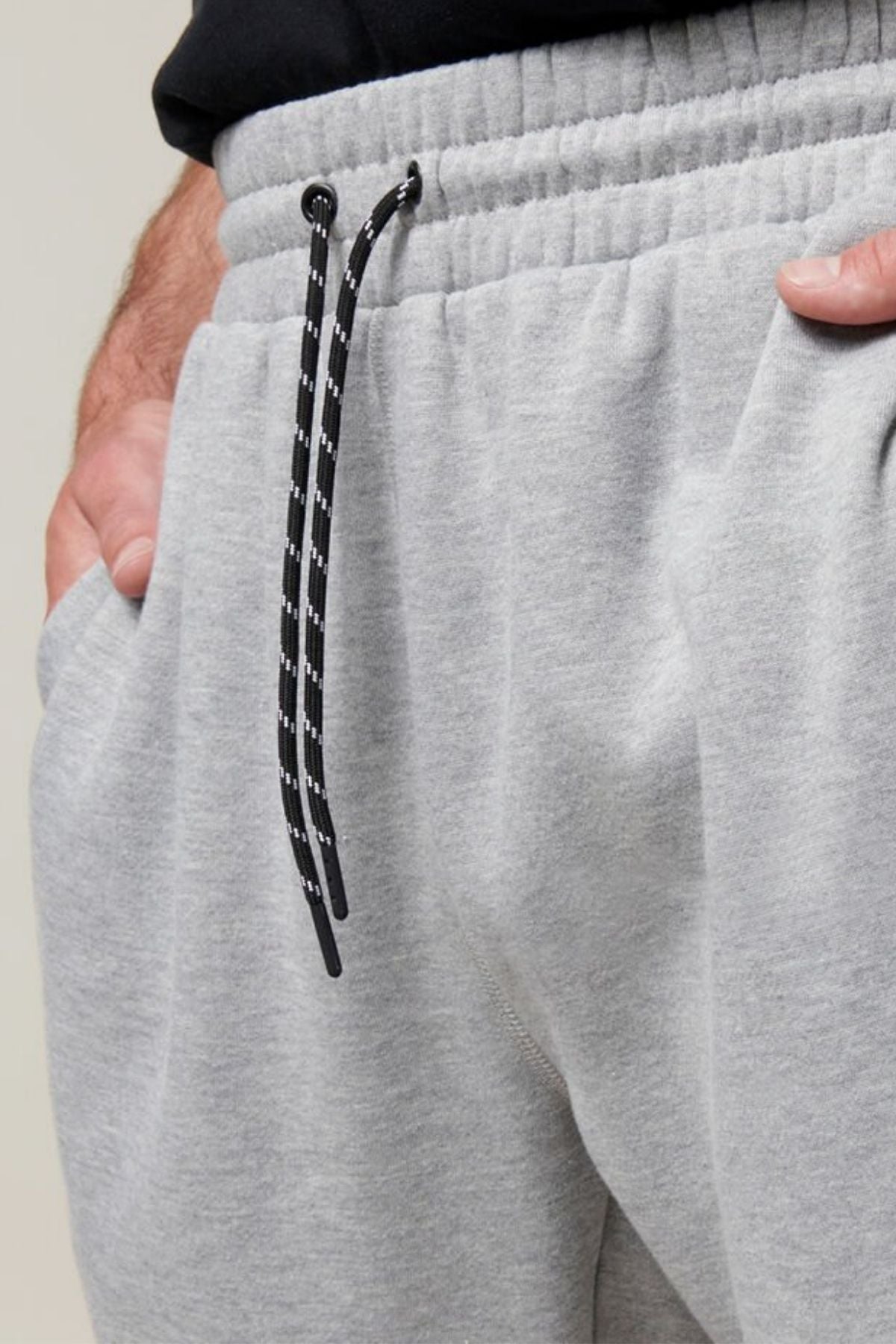A close-up of a person wearing light gray Hard Yakka Xtreme Jogger pants, showcasing the elastic waistband with a black and white drawstring. The person's right hand is partially visible, tucked into the pants' pocket. These joggers feature reinforced knee patches and soft fleece lining. The background is a neutral, light gray color.