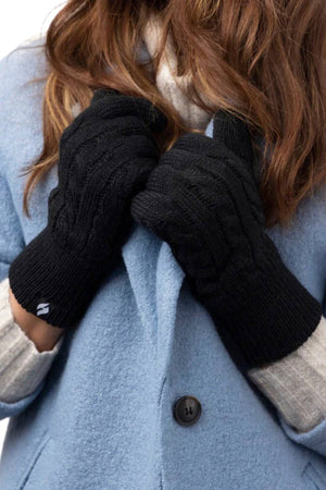A person with long brown hair wears a light blue coat, gray sweater sleeves, and black Heat Holders Willow Ladies Thermal Gloves with a small white logo, holding their hands together near their chest.