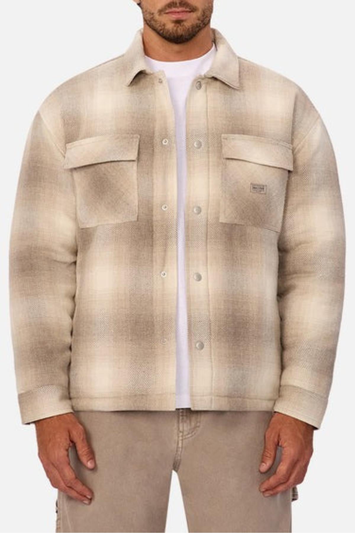 A man wears the Industrie The Highland Jacket in beige and brown plaid with two chest pockets over a white shirt, paired with light brown pants. His hands rest at his sides, exuding readiness for outdoor adventure.