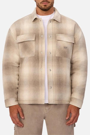 A man wears the Industrie The Highland Jacket in beige and brown plaid with two chest pockets over a white shirt, paired with light brown pants. His hands rest at his sides, exuding readiness for outdoor adventure.