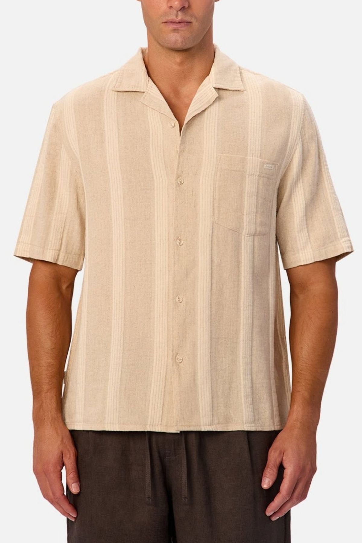 Wearing the Industrie The Ardenza Short Sleeve Shirt—a short sleeve, vertical striped shirt with a camp collar and chest pocket—a man pairs it with dark brown pants and stands against a plain light background.