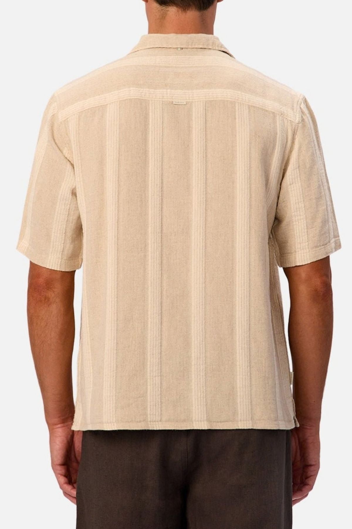 A man is shown from the back wearing the Industrie The Ardenza Short Sleeve Shirt, which features a beige color, vertical stripes, short sleeves, and a camp collar. He is also wearing dark brown pants. The background is plain and light-colored.