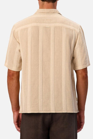 A man is shown from the back wearing the Industrie The Ardenza Short Sleeve Shirt, which features a beige color, vertical stripes, short sleeves, and a camp collar. He is also wearing dark brown pants. The background is plain and light-colored.
