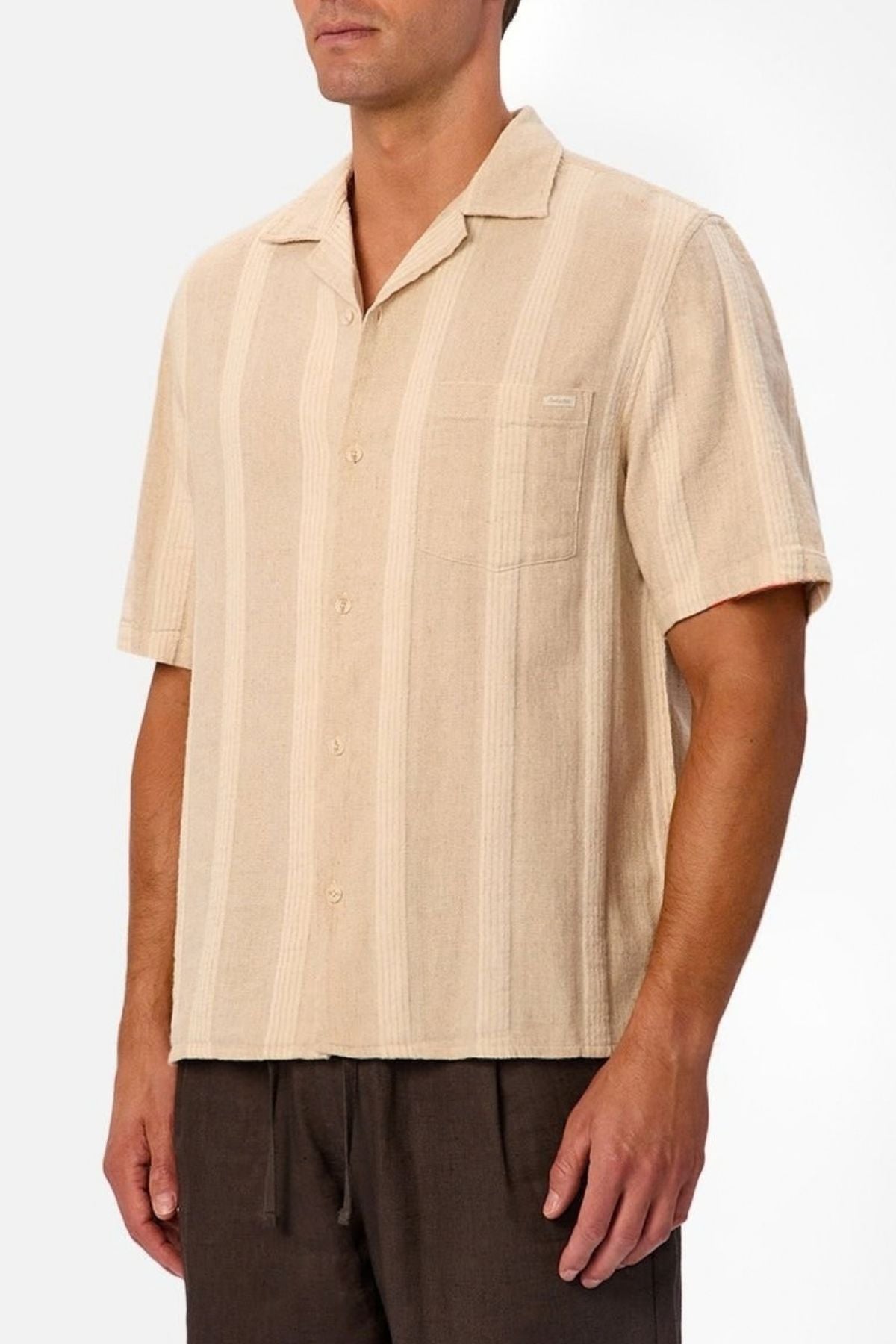 A man stands against a plain white background wearing the Industrie The Ardenza Short Sleeve Shirt—a beige shirt with vertical stripes, a camp collar, and a chest pocket—paired with dark brown pants.