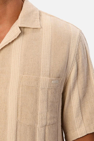 A close-up of the Industrie The Ardenza Short Sleeve Shirt in beige, showcasing its textured vertical stripe pattern, camp collar, and chest pocket. The shirt is worn by a person whose face is not visible.
