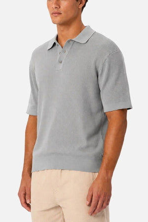 A man stands against a plain white background wearing the Industrie The Bastien Polo—a light gray, short-sleeve shirt crafted from textured fabric with a classic collar.