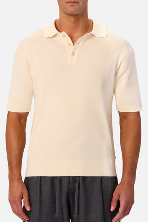 A man wears the cream-colored Industrie The Bastien Polo, which features textured fabric and a classic three-button collar, paired with dark gray pants, standing against a plain light background.