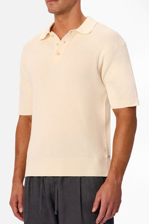 A man wears the Industrie The Bastien Polo in light beige, designed with a classic collar, buttons, and crafted from textured fabric, paired with loose dark gray pants. The background is plain white.