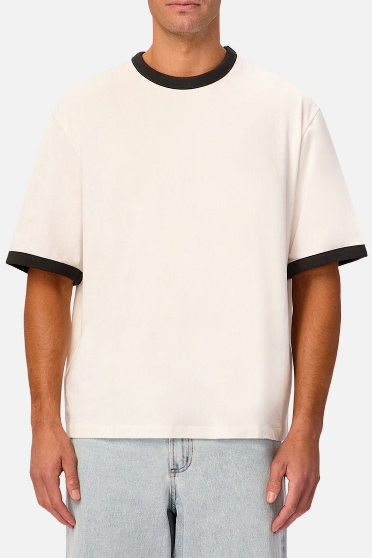 A person wearing the Industrie The Hudson Ringer Tee, a loose-fitting short-sleeve shirt featuring black trim on the classic crew neck and sleeves, paired with light blue jeans and standing against a plain light background.