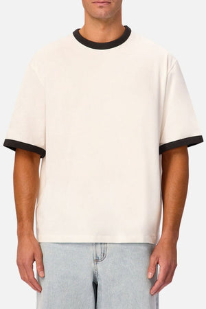 A person wearing the Industrie The Hudson Ringer Tee, a loose-fitting short-sleeve shirt featuring black trim on the classic crew neck and sleeves, paired with light blue jeans and standing against a plain light background.