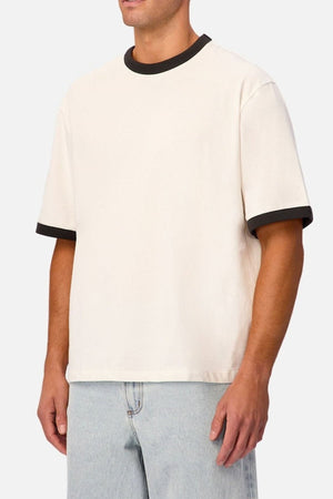 A person wearing the Industrie The Hudson Ringer Tee—a short-sleeve, off-white shirt featuring black trim on the classic crew neckline and sleeves—paired with light blue jeans, stands against a plain light background.