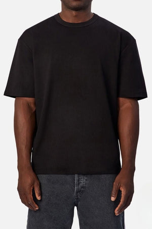 Wearing the Industrie Luxe Del Sur Tee—a relaxed-fit, 100% cotton black t-shirt—and dark gray jeans, a person stands against a white background with arms at their sides.