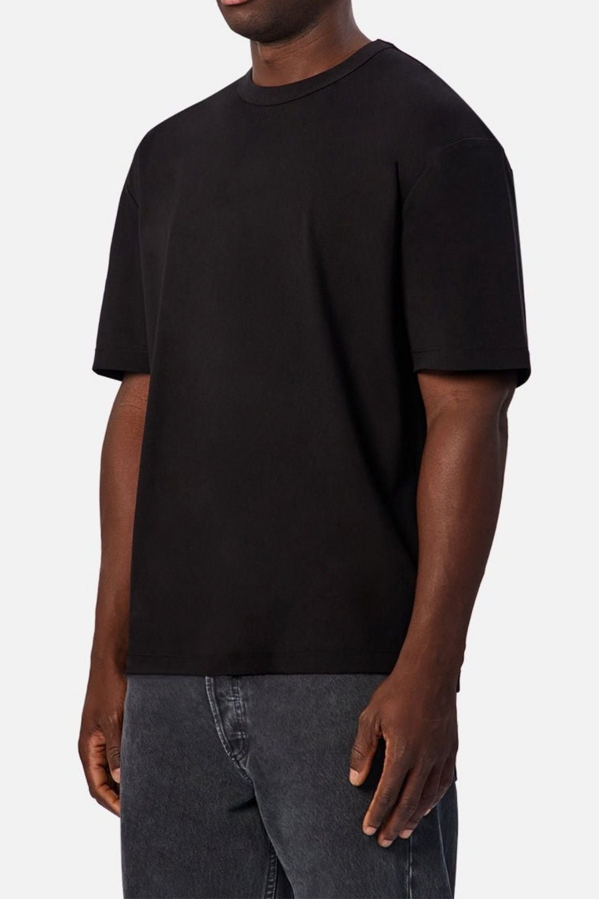 A person stands against a light gray background wearing dark gray jeans and the Industrie Luxe Del Sur Tee, a relaxed-fit style made from 100% cotton. The image is cropped from the shoulders to the upper thighs.