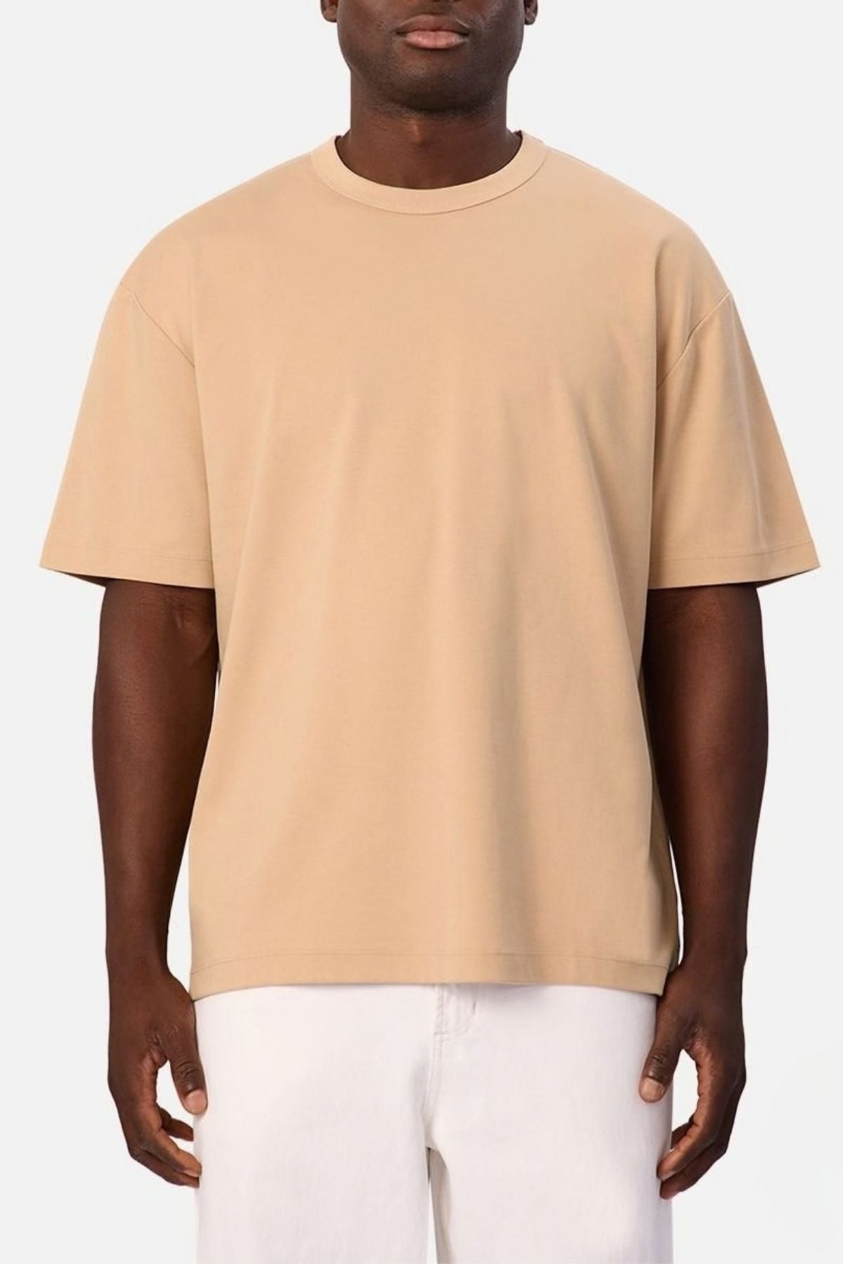 Wearing the Industrie Luxe Del Sur Tee—a relaxed-fit T-shirt made from 100% cotton in plain beige—a person stands against a light background. Their white pants contrast with the tee, and their arms hang loosely at their sides, while their face is partially visible.