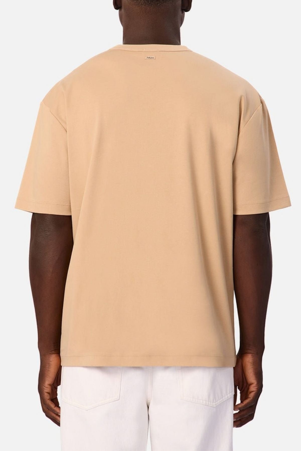 A person is shown from the back against a plain light background, wearing the Industrie Luxe Del Sur Tee in a relaxed-fit beige short-sleeve style paired with white pants. The T-shirt is crafted from 100% cotton for exceptional comfort and breathability.