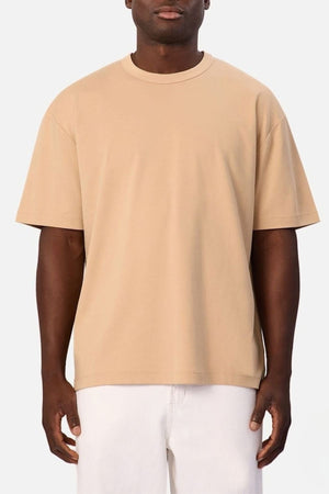 Wearing the Industrie Luxe Del Sur Tee—a relaxed-fit T-shirt made from 100% cotton in plain beige—a person stands against a light background. Their white pants contrast with the tee, and their arms hang loosely at their sides, while their face is partially visible.