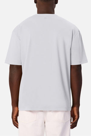 A person is seen from the back against a plain white background, wearing the Industrie Luxe Del Sur Tee—a relaxed-fit, light gray T-shirt made from 100% cotton—paired with light-colored pants.