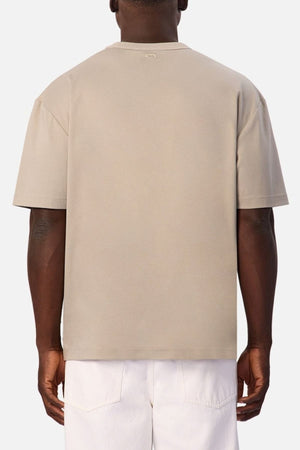 A person is shown from the back wearing the beige Industrie Luxe Del Sur Tee, a 100% cotton relaxed-fit top, paired with white pants and standing against a plain light background.