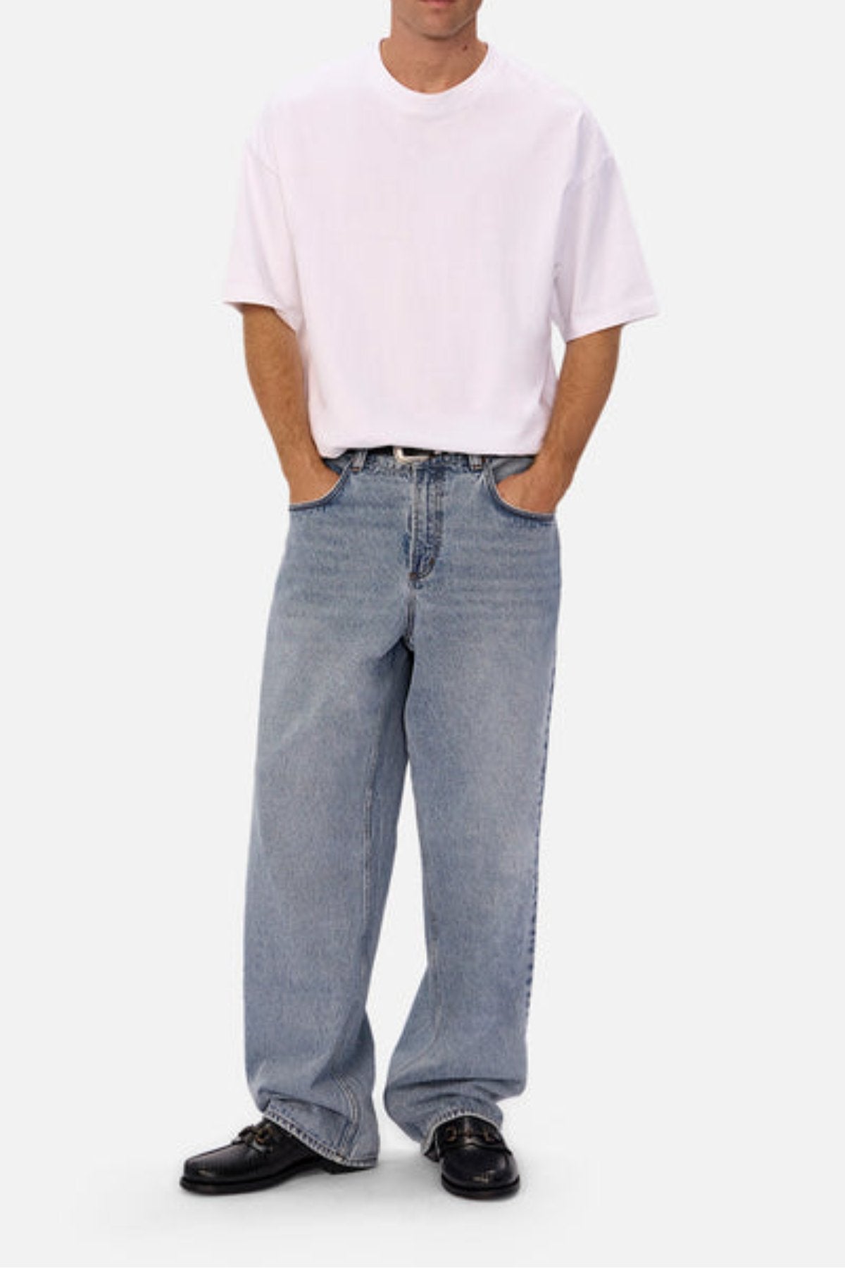 A person stands with hands in pockets, wearing a plain white T-shirt, black shoes, and Industrie The 80's Jean in a relaxed fit light blue durable denim. The background is plain and light-colored; the face isn't fully visible.