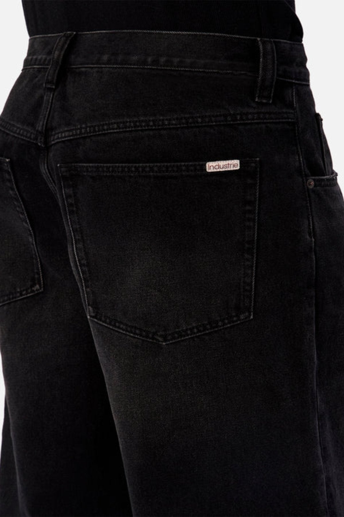 Close-up of the back of a person wearing Industrie The 80's Jean in black denim, featuring a visible back pocket and a small "Industrie" label above it. The person pairs the jeans with a black top.