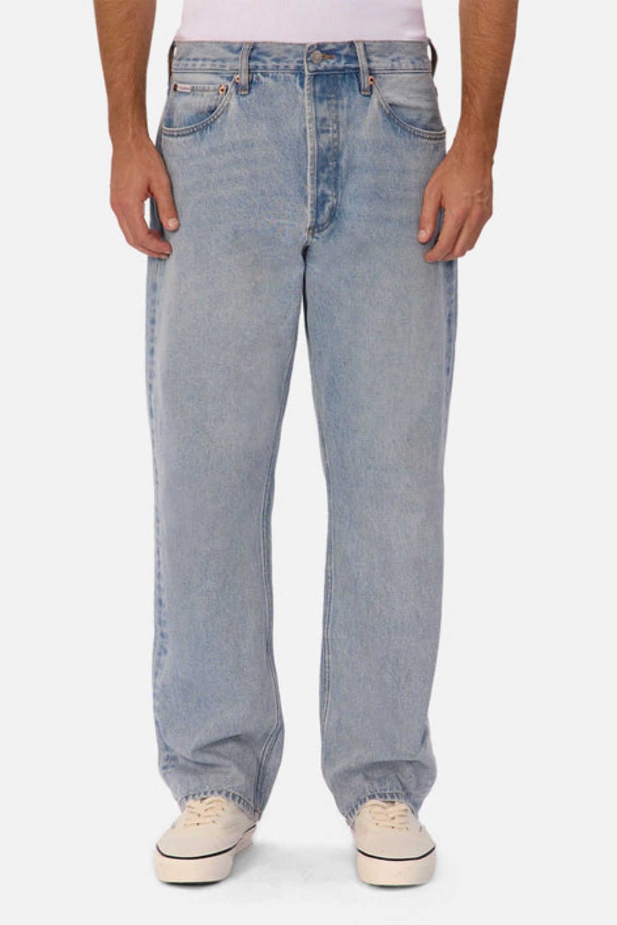 Someone wearing Industrie The 90's Jean in vintage washed light blue, paired with a white shirt and white sneakers, stands facing forward against a plain white background.