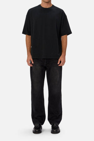 A person stands against a plain background, wearing a loose black t-shirt and Industrie The 90's Jean—relaxed, straight-leg, vintage-washed denim. Their hands are at their sides and their face isn’t visible.