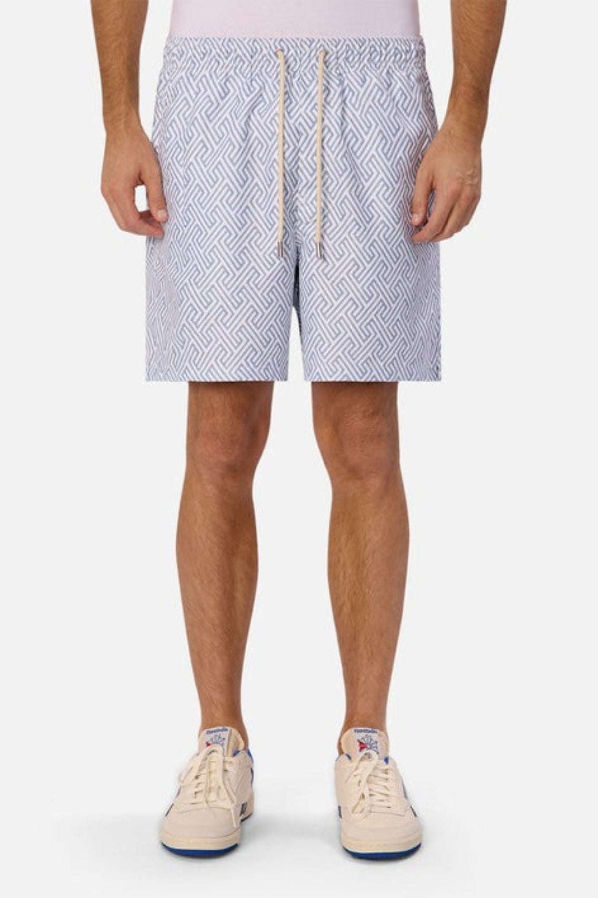Someone is standing against a plain white background wearing Industrie The Alvarado Swim Shorts and white sneakers. The swim shorts, celebrated for their intricate geometric design and regular tailored fit, pair well with the sneakers featuring blue accents. The person's upper body is not visible.