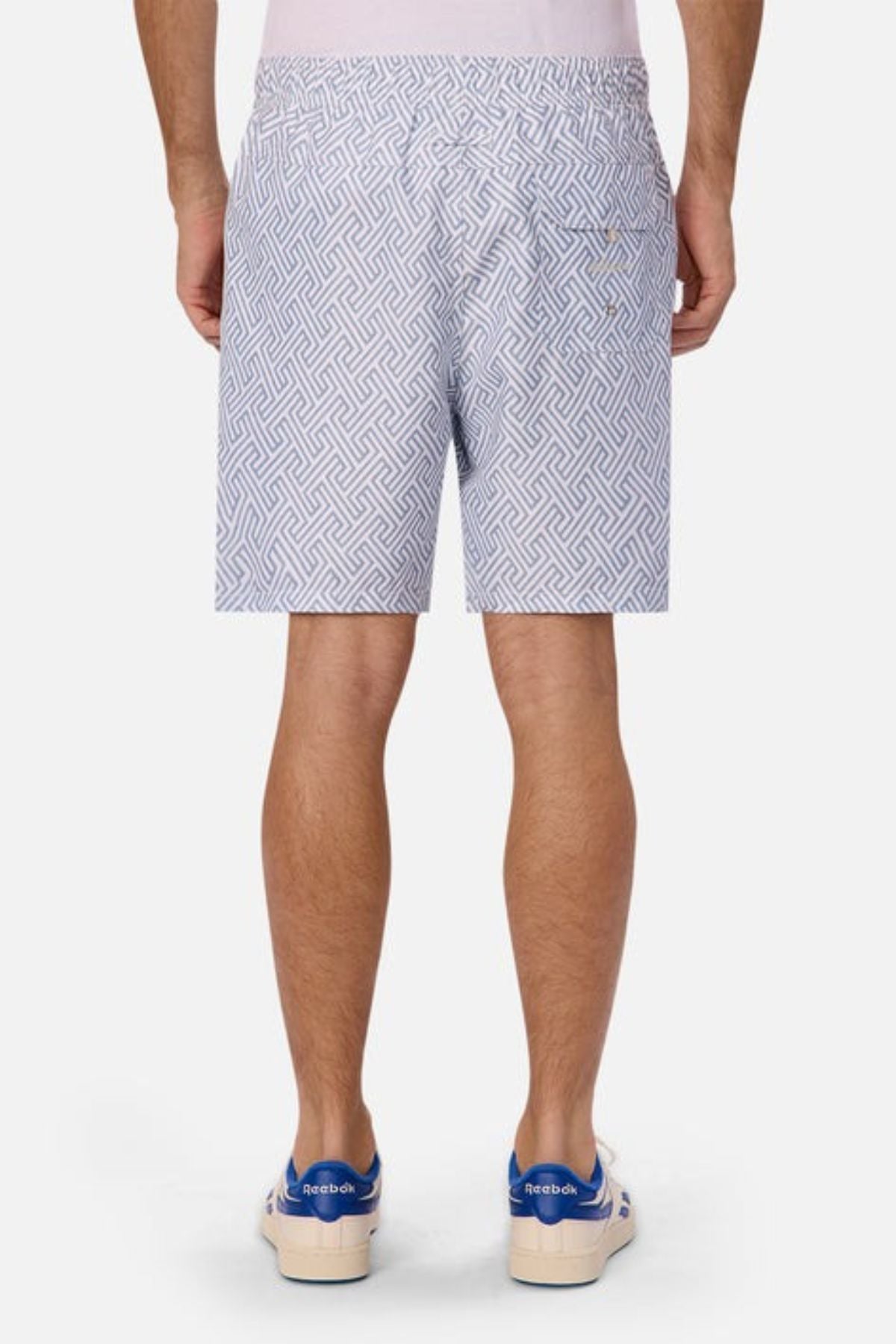 Rear view of a person wearing Industrie The Alvarado Swim Short with a tailored regular fit and white sneakers featuring blue accents, standing against a plain background.