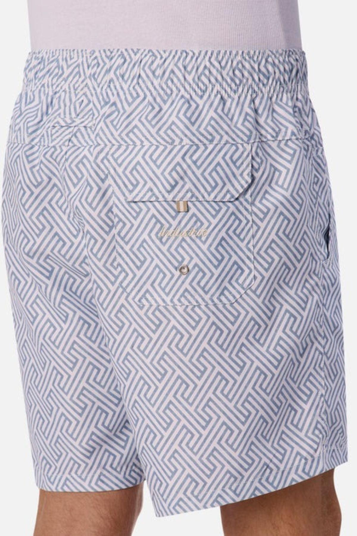 A person wearing the Industrie The Alvarado Swim Short, showcasing a white and light blue patterned fabric with an elastic waistband. These tailored, regular-fit shorts feature a geometric design and include a back pocket with button closure, set against a plain background.