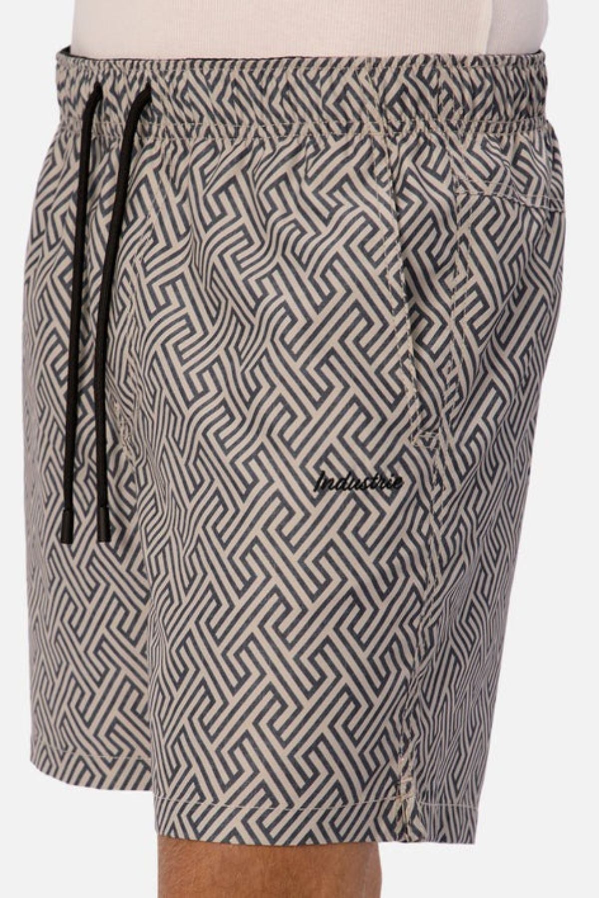A person wearing the Industrie The Alvarado Swim Short, which showcases a geometric maze-like design in gray and black. These quick-drying shorts include a black drawstring and have a small logo near the pocket, making them perfect for aquatic leisure.