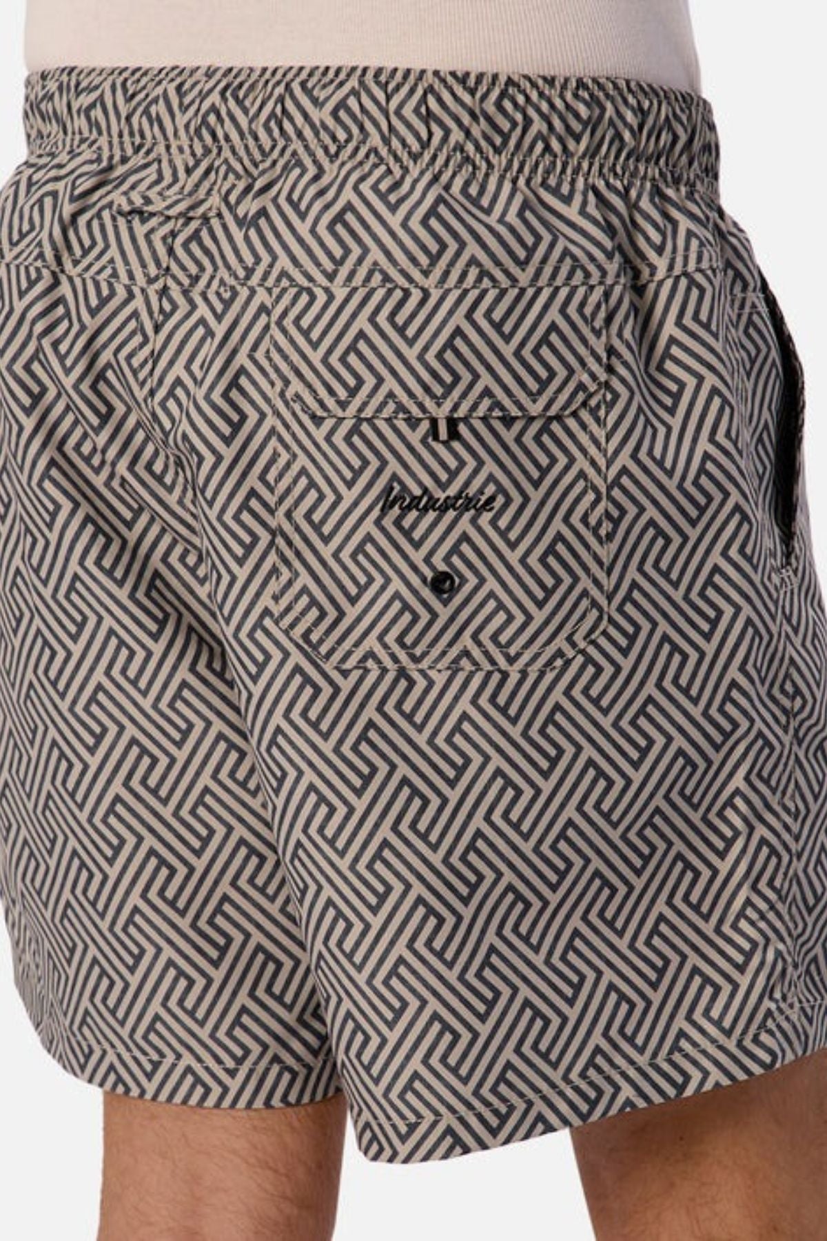 A close-up of someone wearing the Industrie The Alvarado Swim Short, showcasing its geometric pattern in black and gray. Ideal for water activities, these quick-drying shorts come with an elastic waistband, a back pocket secured by a small button, and proudly display the brand's logo.