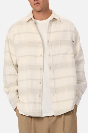 A man wears the Industrie The Arizona Long Sleeve Shirt, a cream classic-fit striped button-up, layered over a white t-shirt with tan pants, standing with hands in pockets against a plain background.