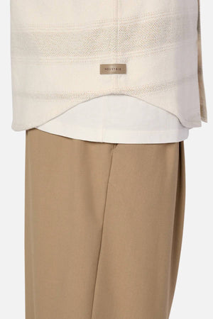 A close-up shows a person wearing the Industrie The Arizona Long Sleeve Shirt in cream, made from 100% cotton, layered over a white top with loose beige pants. A small metal "INDUSTRIE" tag is attached at the hem.