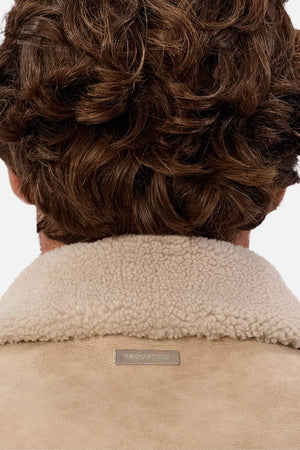 A close-up of a person with wavy brown hair wearing the Industrie The Artem Flight Jacket in tan PU leather, featuring a Sherpa-look collar and a small rectangular label at the neckline.