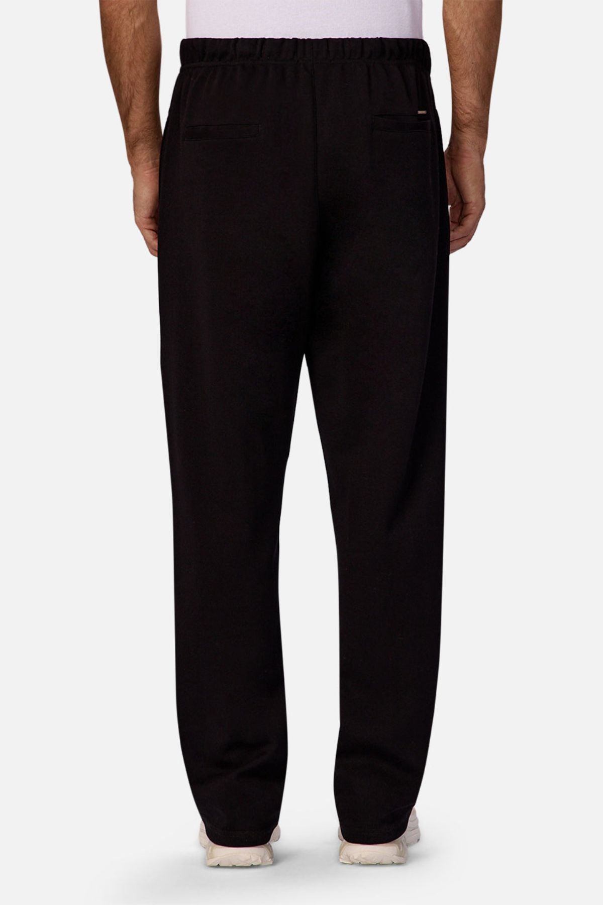 A person is shown from the back wearing Industrie The Astoria Lux Tech Trackpant in black with an elastic waistband, two welt back pockets, white sneakers, and a partially visible white shirt, blending casual style and premium comfort.