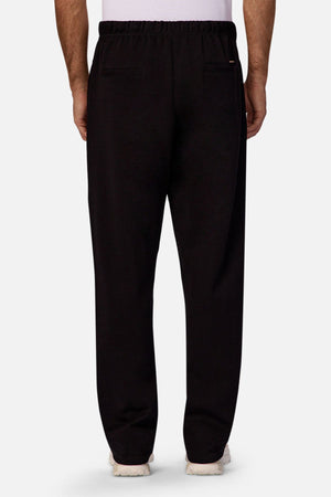 A person is shown from the back wearing Industrie The Astoria Lux Tech Trackpant in black with an elastic waistband, two welt back pockets, white sneakers, and a partially visible white shirt, blending casual style and premium comfort.