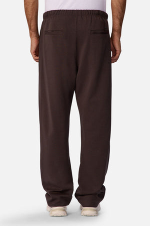 Someone wearing the Industrie The Astoria Lux Tech Trackpant in dark brown, featuring an elastic waistband and two back pockets, is shown from the back against a plain light background for a casual style look.