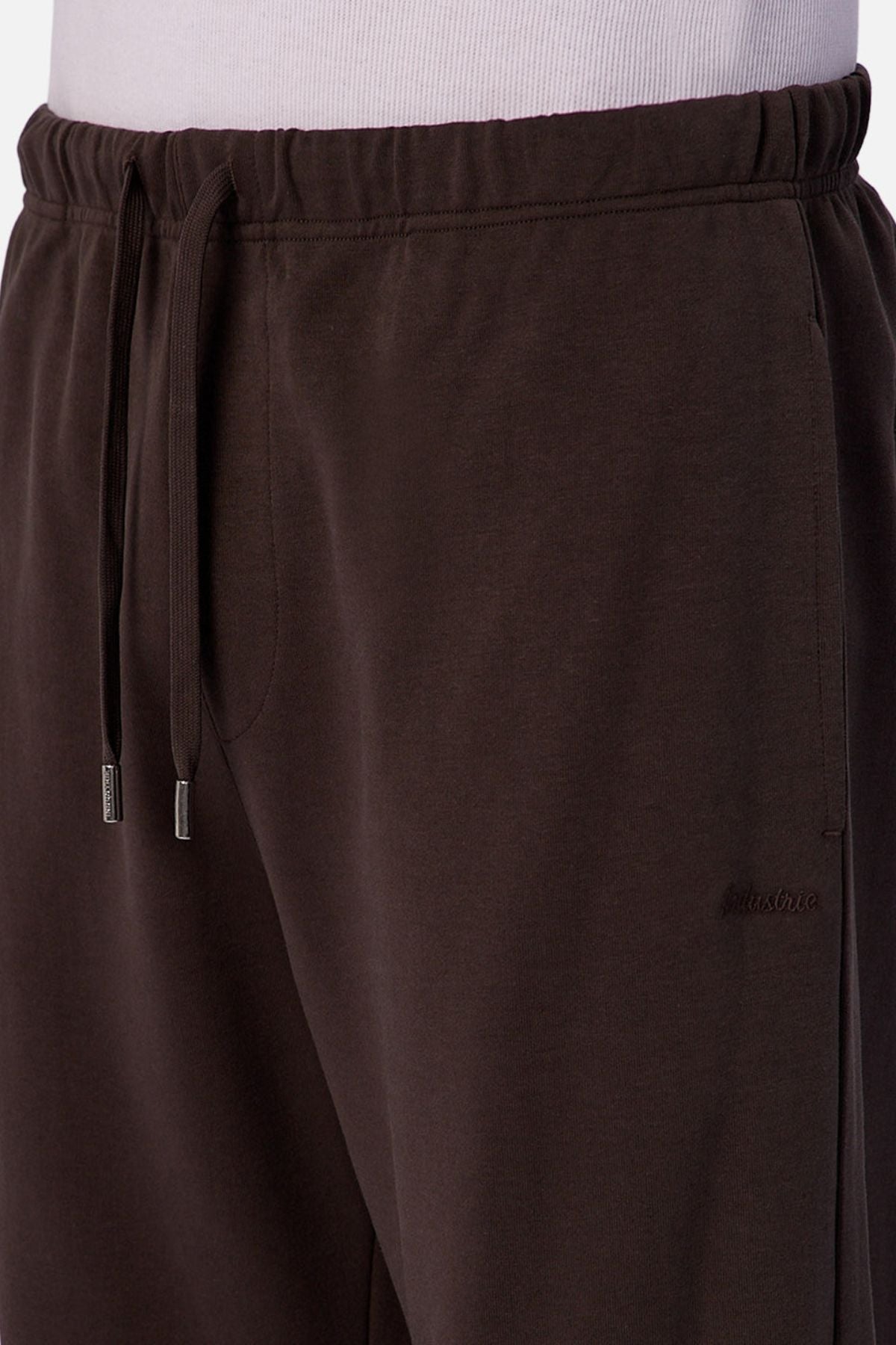 Close-up of a person wearing Industrie The Astoria Lux Tech Trackpant in dark brown, featuring a drawstring waistband, side pockets, and a subtle embroidered logo on the left thigh. Paired with a white shirt for casual, premium comfort.