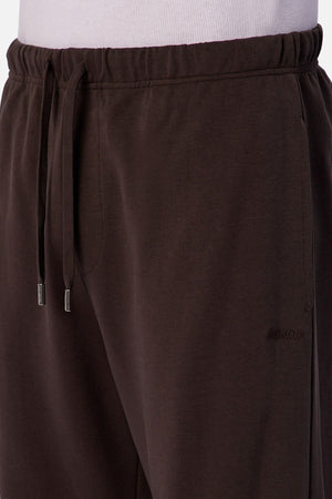 Close-up of a person wearing Industrie The Astoria Lux Tech Trackpant in dark brown, featuring a drawstring waistband, side pockets, and a subtle embroidered logo on the left thigh. Paired with a white shirt for casual, premium comfort.