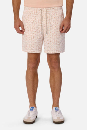 A person wearing Industrie The Avila Swim Short, which features a peach color with a subtle geometric pattern and a relaxed fit, paired with a light peach shirt and white sneakers accented in blue, stands against a plain background. Only the lower half of their body is visible.