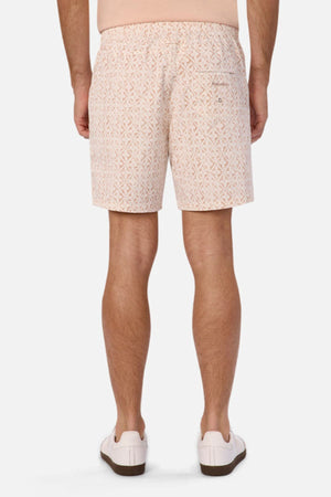 A person is wearing The Avila Swim Short in a relaxed fit with a pattern in beige, paired with white sneakers, standing with their back to the camera against a plain background.