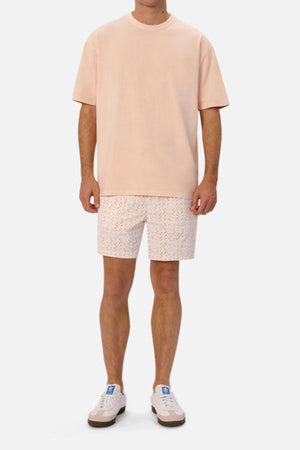 A person wearing a light peach-colored oversized T-shirt with matching Industrie The Avila Swim Short stands against a plain background. They are also wearing white sneakers with blue accents. The overall look is casual and summery, perfect for quick-drying comfort by the poolside.