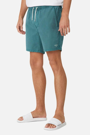 A person in Industrie The Bahama Angler Short, teal swimming shorts with an elastic waistband and drawstring, stands in a partial side view wearing white slide sandals, all on a crisp white background.