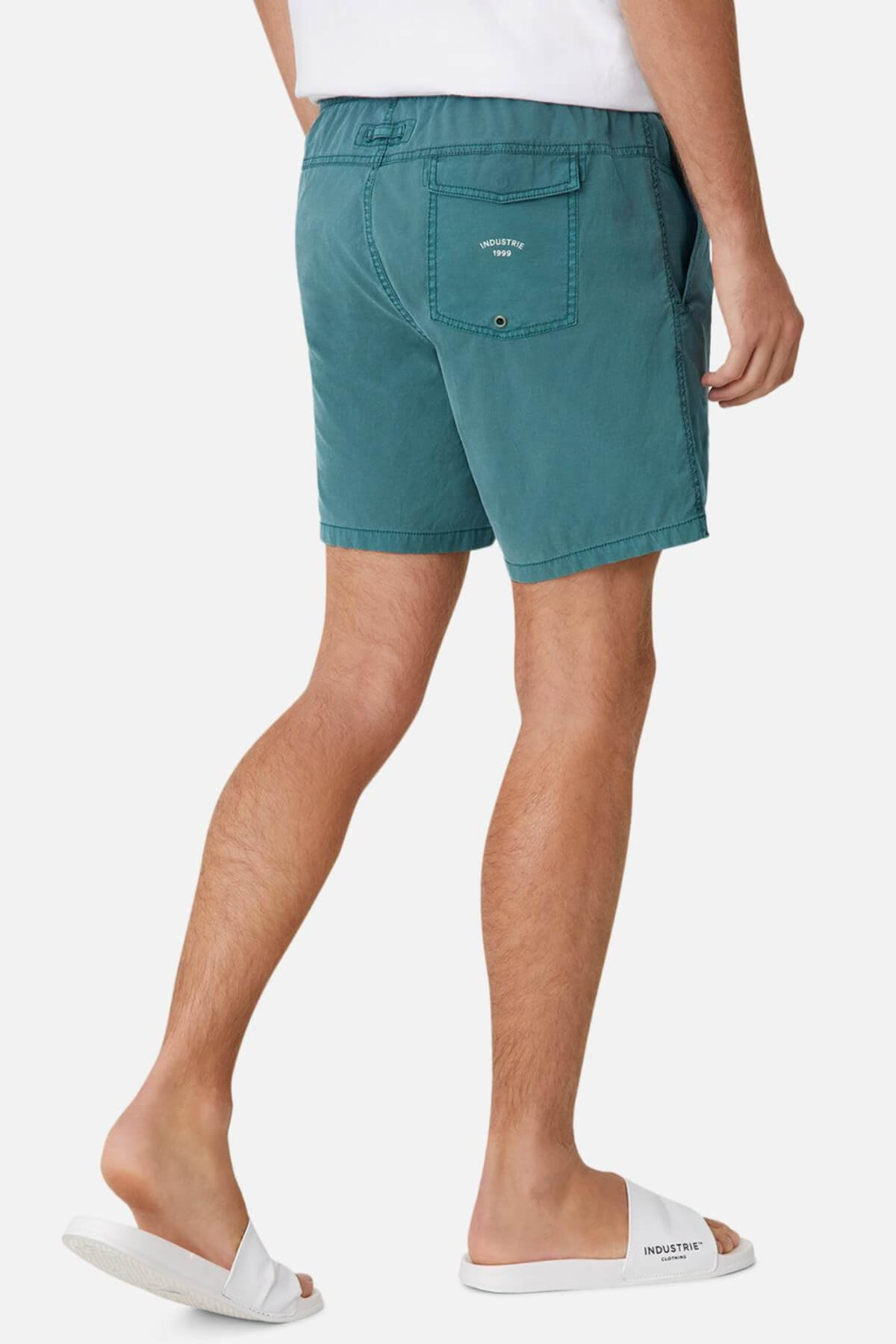 Someone is wearing Industrie The Bahama Angler Short in teal with an elastic waist, back pocket with a button, visible stitching, and white slide sandals. They're standing sideways on a white background, showing only their legs and part of a white shirt.