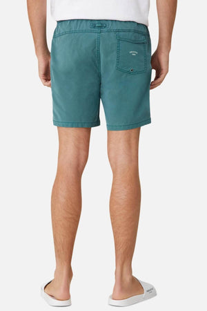 A person is shown from the back wearing Industrie The Bahama Angler Short in teal, featuring an elastic waist and a buttoned back pocket, along with white flip-flops. The background is plain white.