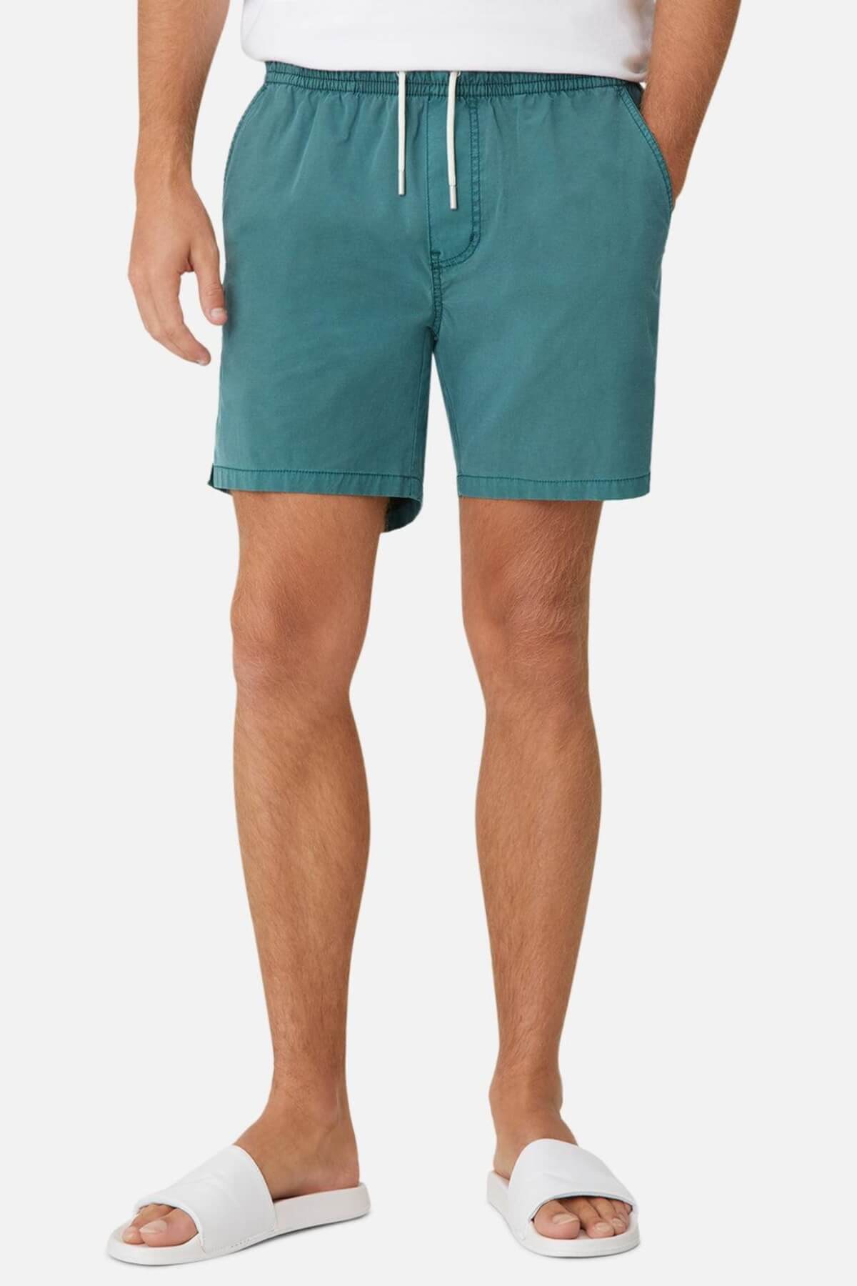 Wearing Industrie The Bahama Angler Shorts in teal, featuring an elastic waist and white drawstring, hands casually in pockets. The outfit is completed with a crisp white shirt and matching slide sandals against a simple backdrop, highlighting the relaxed style.