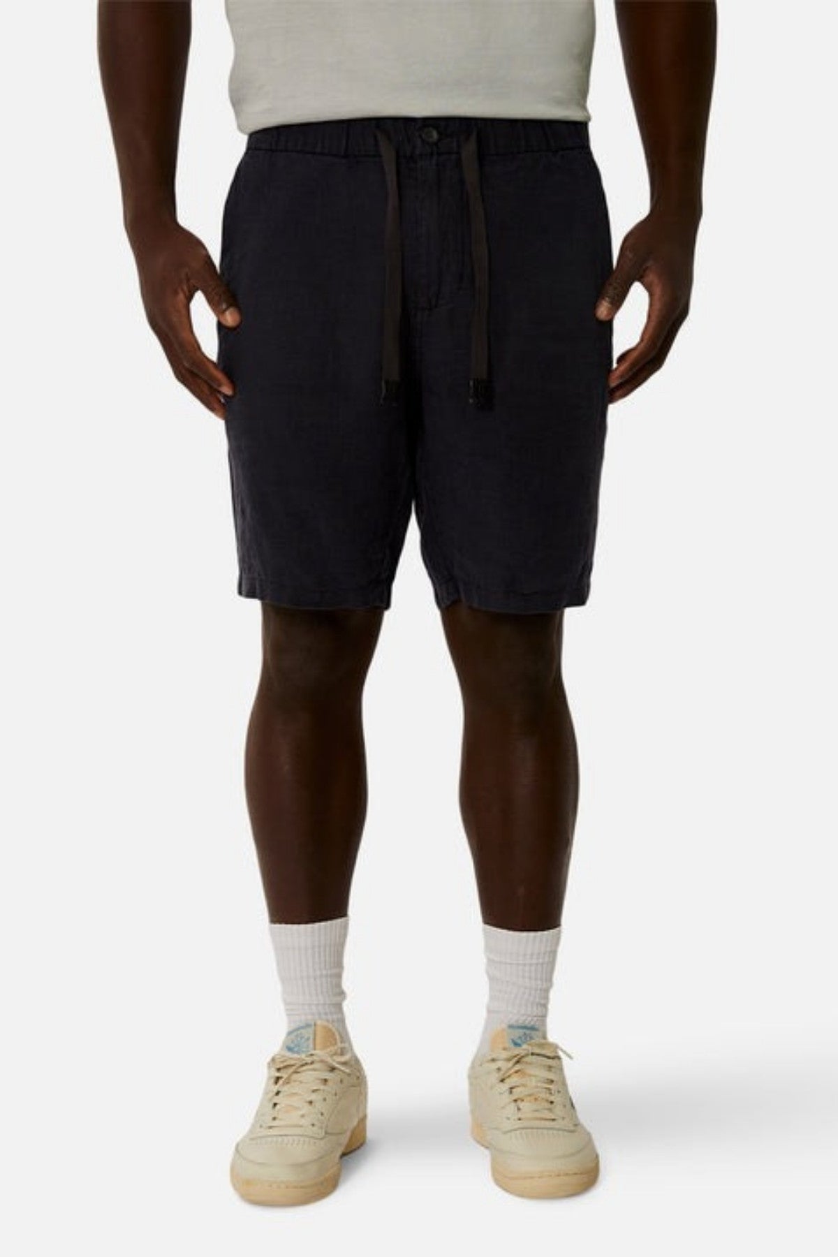 A person wearing a light gray shirt, Industrie The Baller Linen Short in dark blue with a drawstring waist and relaxed fit, and white socks with cream-colored sneakers featuring light blue accents stands against a plain background.